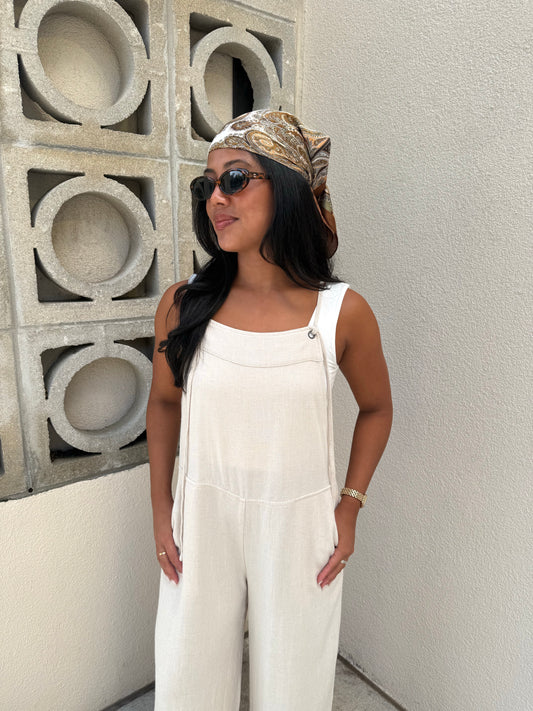 A woman wearing a white sleeveless jumpsuit, a brown and gold paisley headscarf, and sunglasses, standing against a textured beige wall with a concrete block pattern.