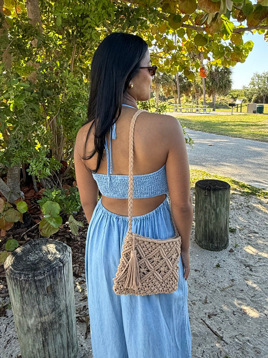 Woman in a blue dress with a macrame purse standing outdoors near trees and a path.