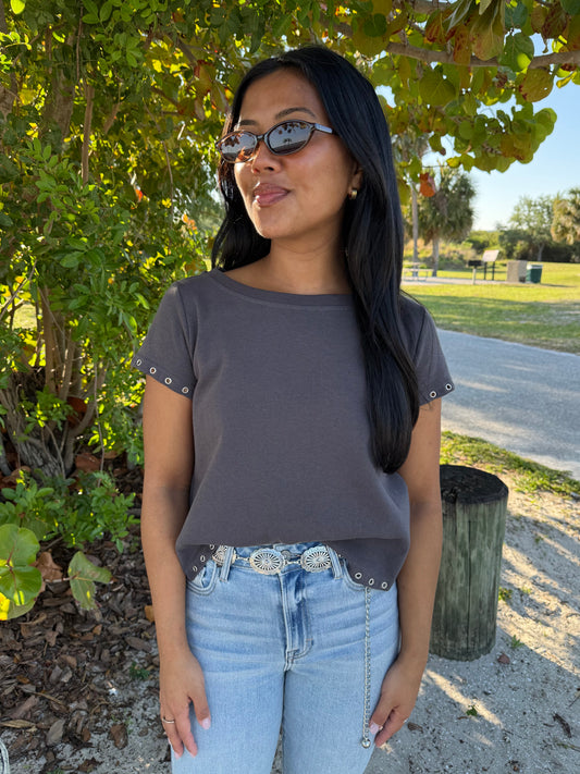 Woman wearing sunglasses and a dark gray top with light blue jeans in an outdoor setting.