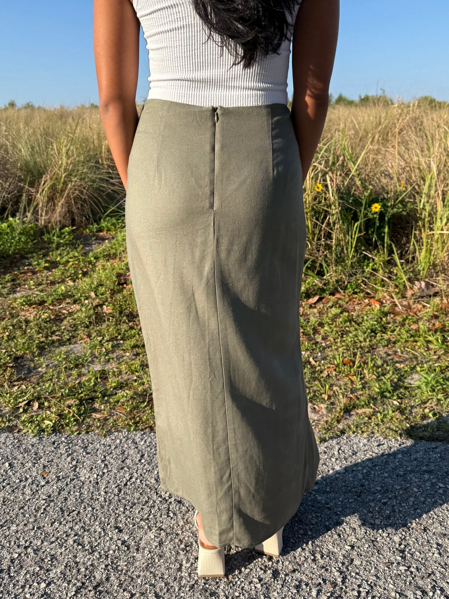 Person wearing a long olive green skirt standing on a gravel path with grass and wildflowers in the background.