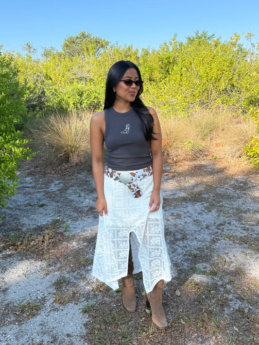 Woman standing outdoors in a natural setting wearing sunglasses, a tank top, and a long white skirt.