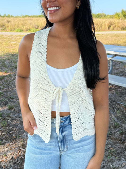 Woman wearing a white crochet vest over a white top and blue jeans outdoors.