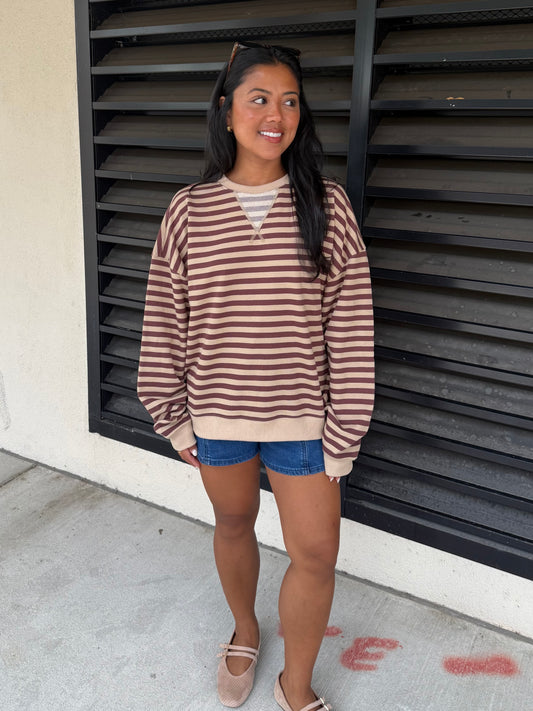 A woman wearing a brown and beige horizontally striped long-sleeve crewneck sweatshirt, denim shorts, and beige flats, standing in front of a black metal louvered door.