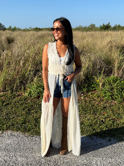 Woman wearing a long white lace dress standing in a natural setting with grass and trees.