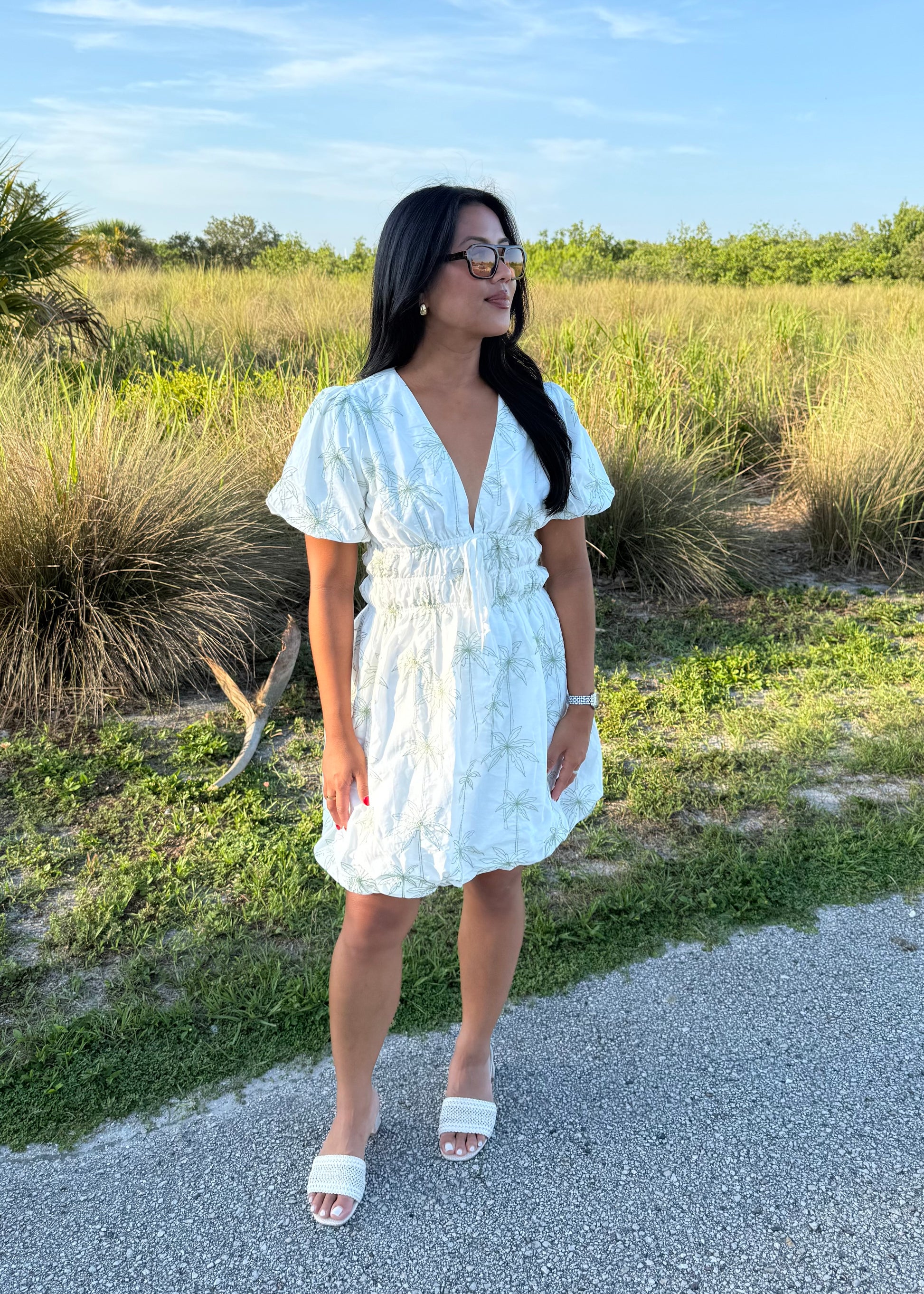 Girl wearing white mini dress with green palm tree print on a grass background.