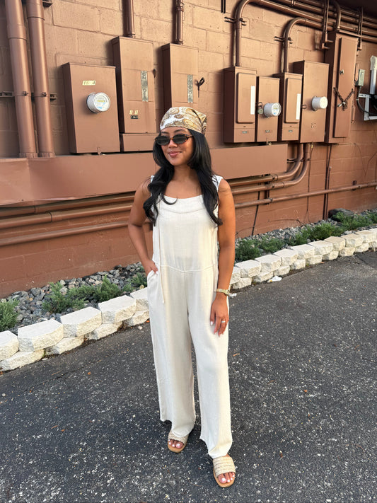 A woman wearing a white sleeveless wide-leg jumpsuit, sunglasses, and a patterned headband stands outdoors in front of a brown utility wall with electrical boxes and pipes.