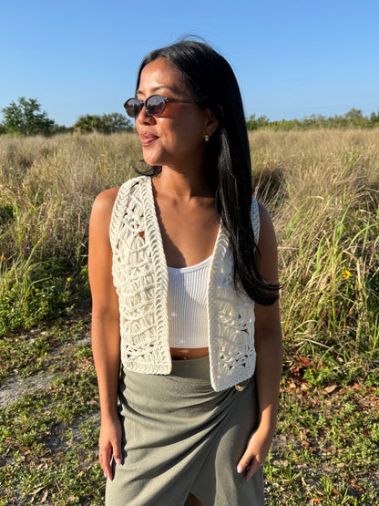 Woman standing in a field wearing sunglasses and a white crochet vest.