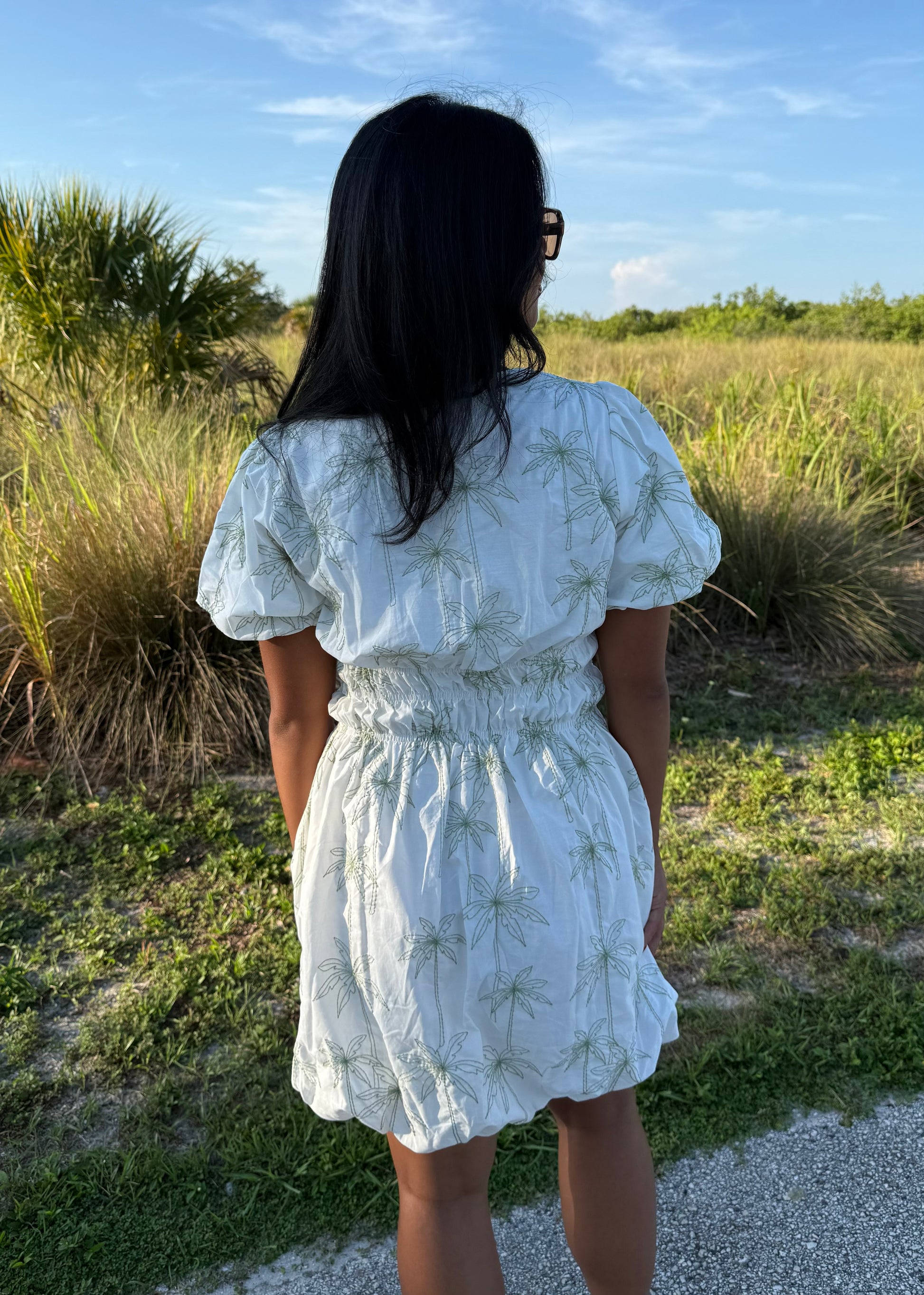 Girl wearing white mini dress with green palm tree print on a grass background.