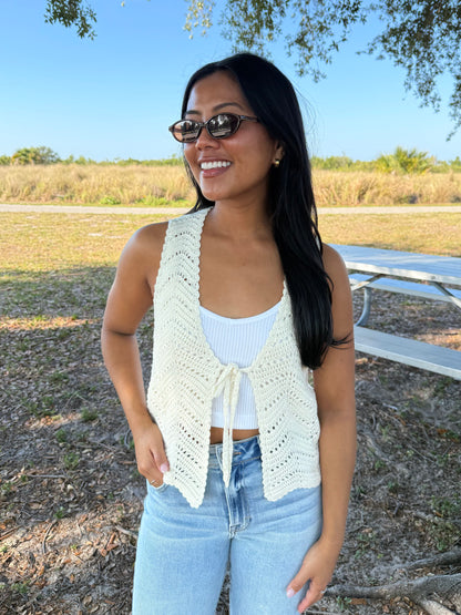 Woman wearing a white crochet vest over a light blue top and jeans, standing outdoors.