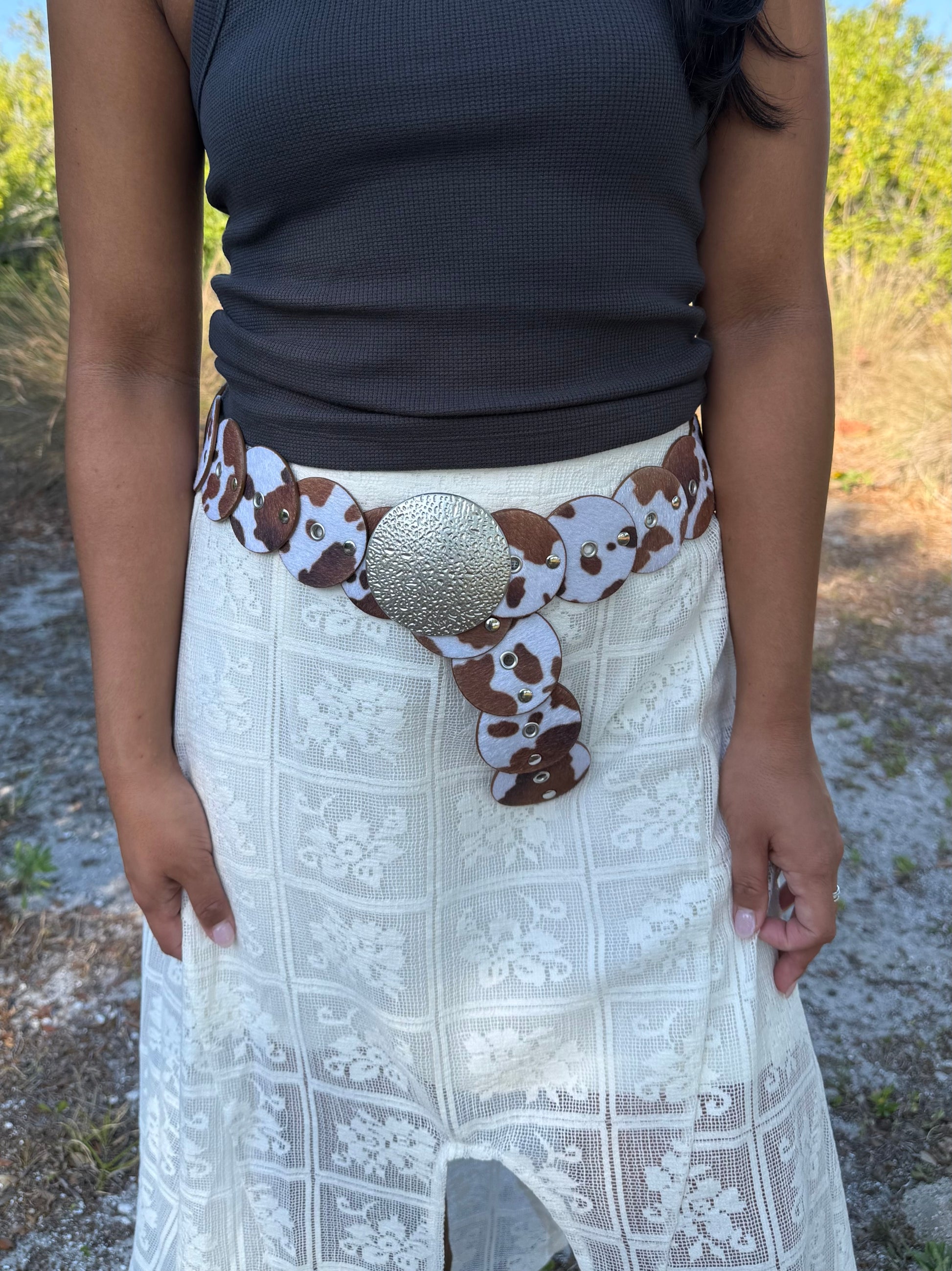 Person wearing a cow print belt with a large round buckle outdoors.