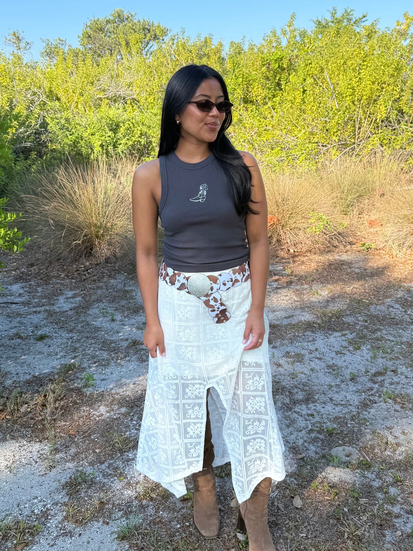 Woman standing outdoors in a natural setting wearing sunglasses, a tank top, and a long white skirt.