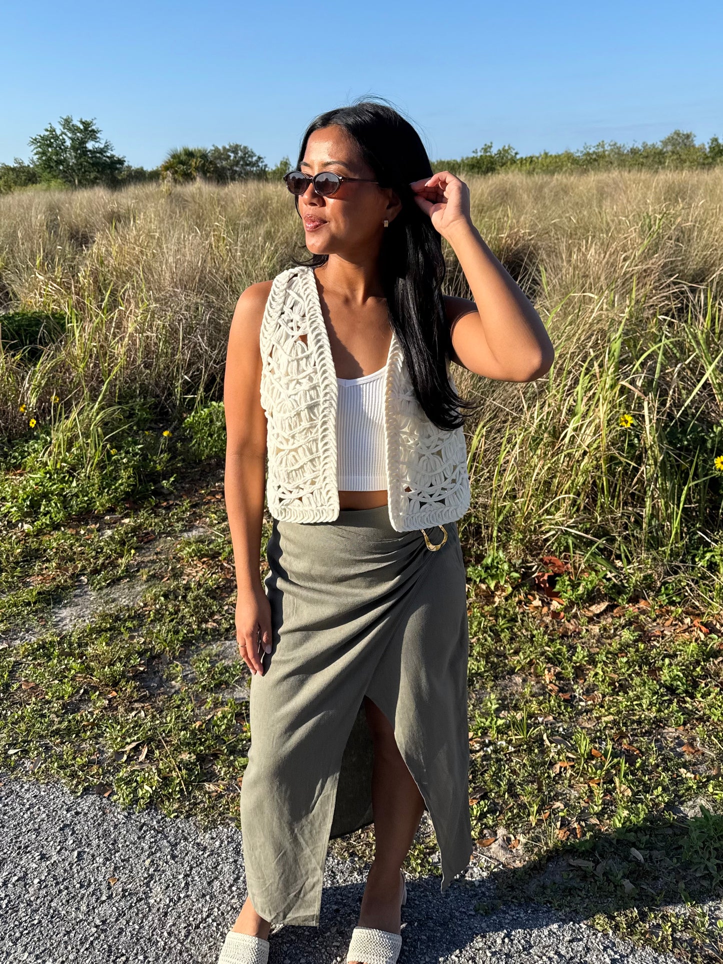 Woman standing in a field wearing a white crochet vest and gray skirt.