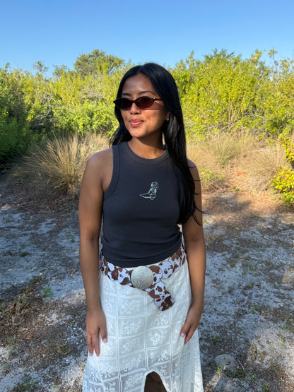 Woman standing outdoors in a natural setting wearing sunglasses and a black tank top with a white skirt.