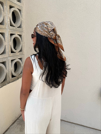 A woman wearing a white sleeveless jumpsuit and a brown patterned headscarf, photographed from behind in an outdoor setting next to a concrete block wall.