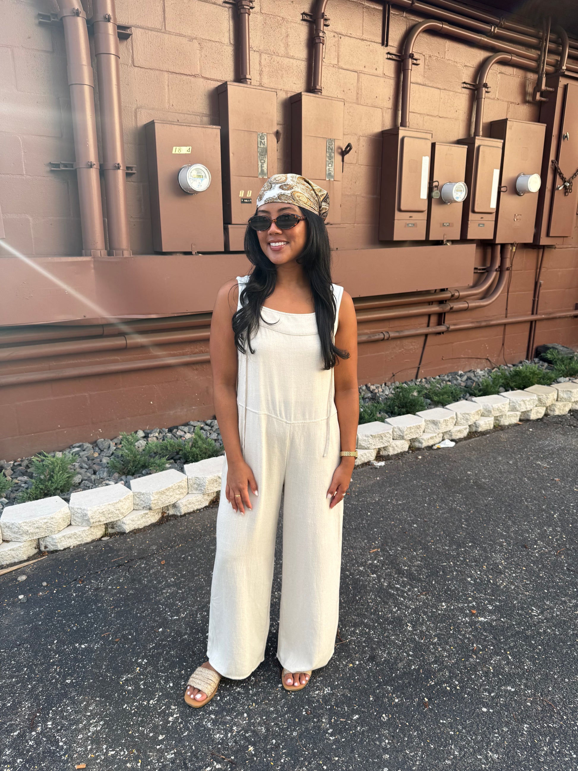 A woman wearing a cream wide-leg jumpsuit, sunglasses, and a patterned headwrap stands outdoors in front of a brown brick wall with utility boxes.