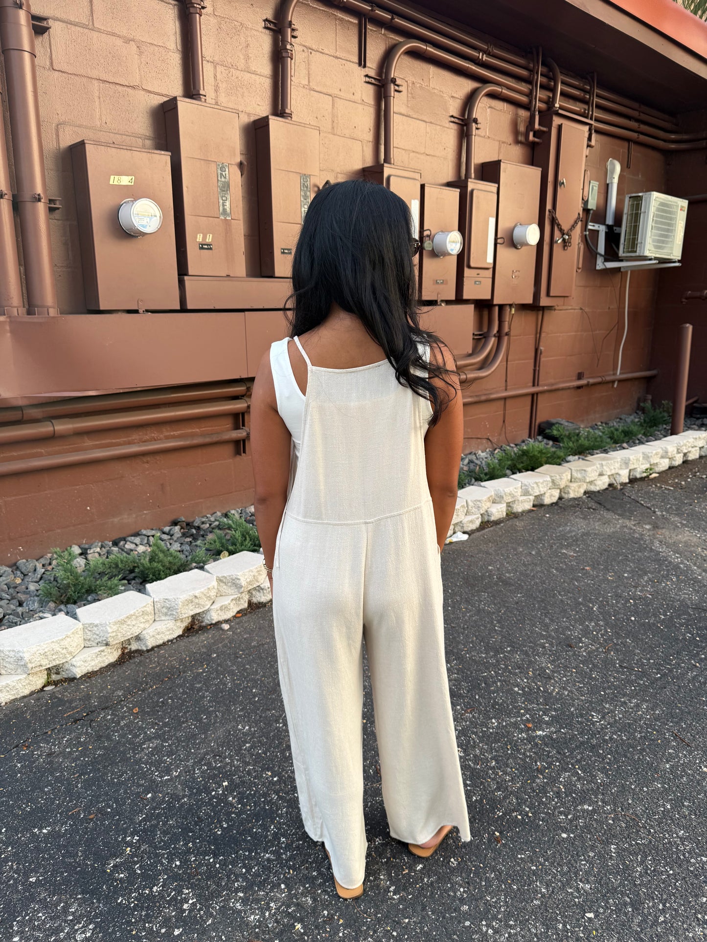 A woman viewed from behind wearing a cream-colored wide-leg jumpsuit, standing on asphalt in front of a brown utility wall with electrical boxes.
