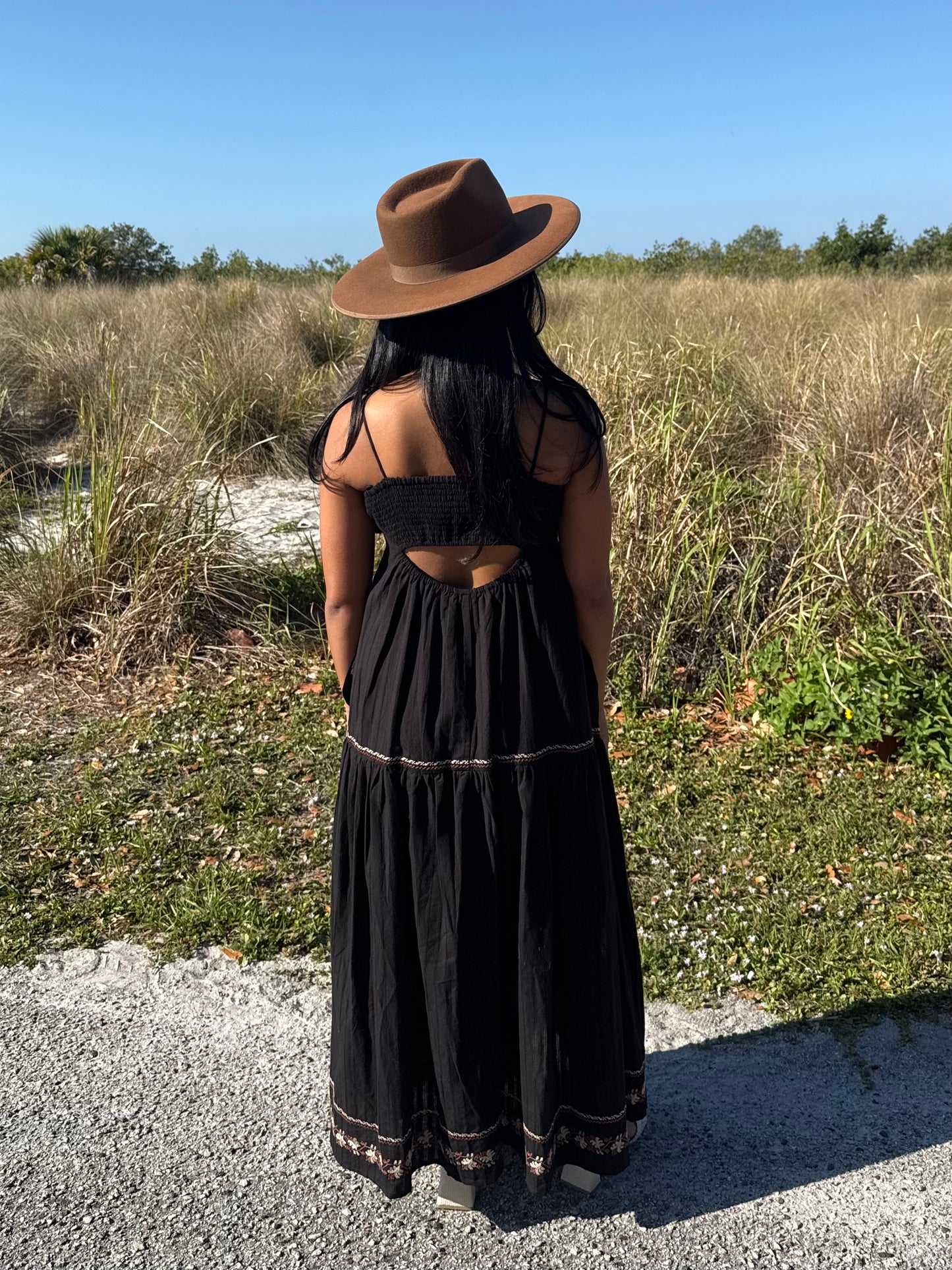 Person wearing a black dress and brown hat standing in a natural setting with grass and clear sky.