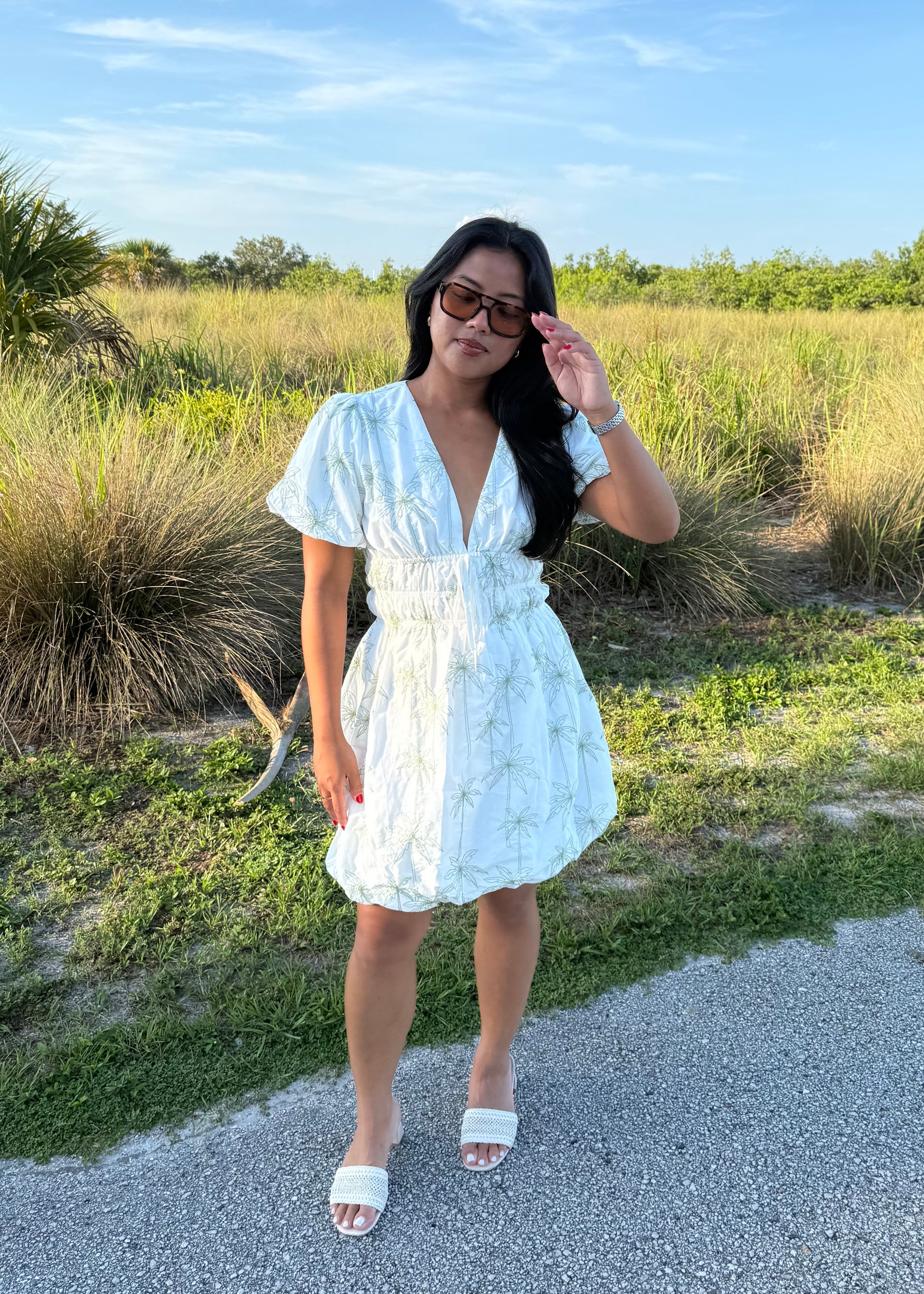 Girl wearing white mini dress with green palm tree print on a grass background.