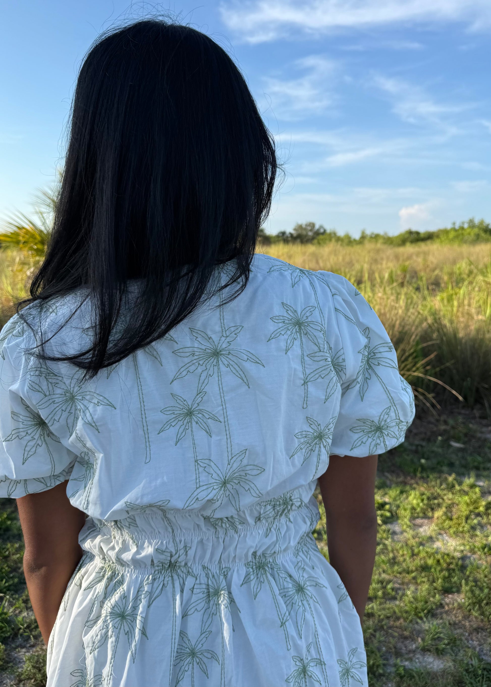 Girl wearing white mini dress with green palm tree print on a grass background.