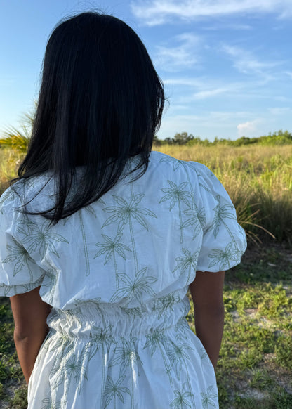 Girl wearing white mini dress with green palm tree print on a grass background.