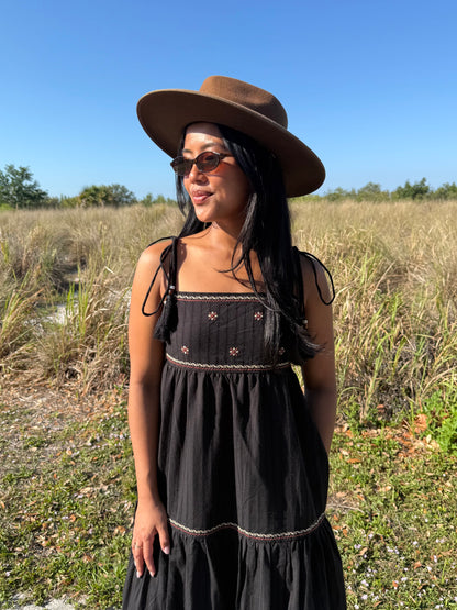 Woman wearing a black dress and brown hat standing in a field with a clear blue sky.