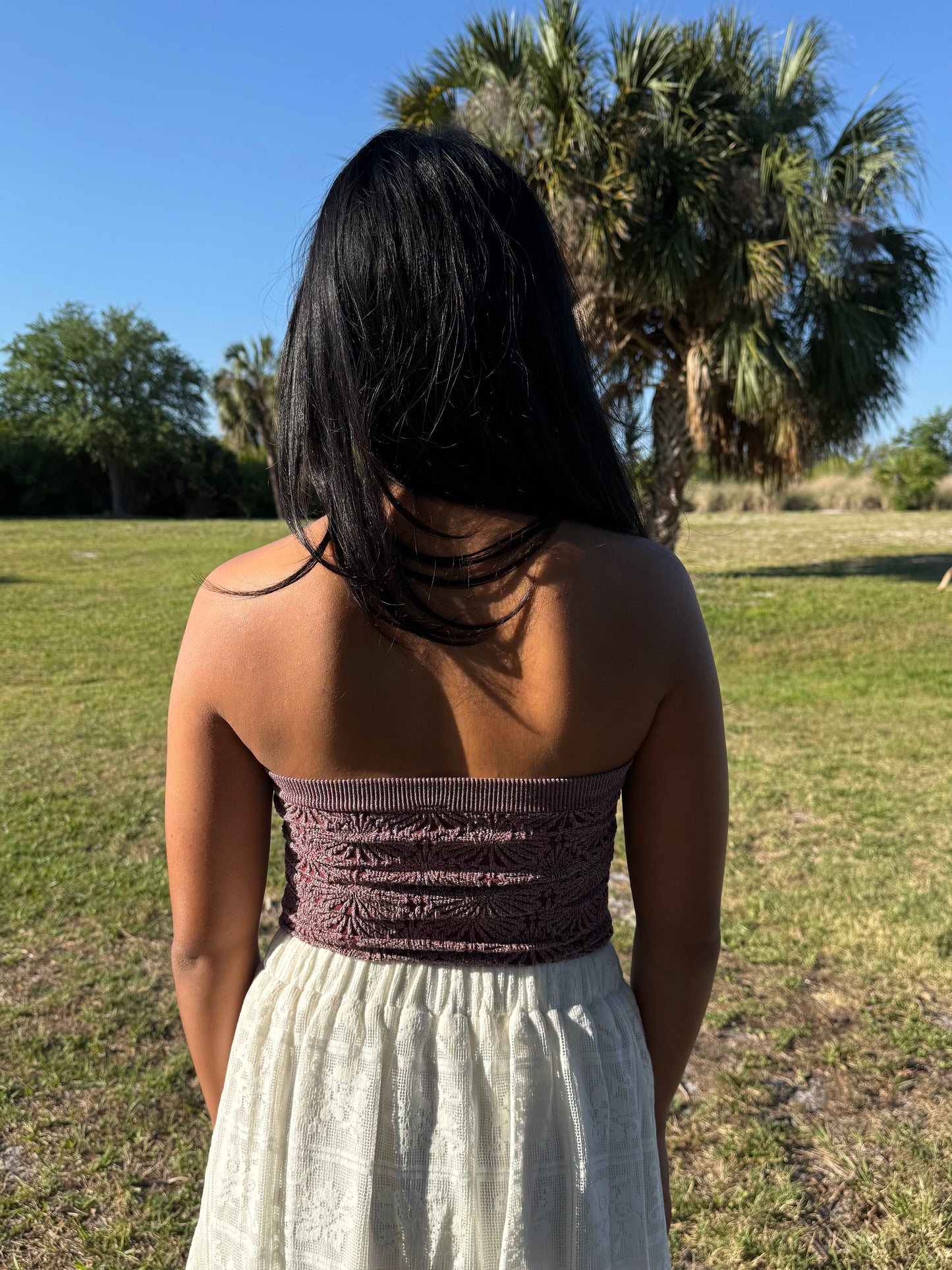 Person wearing a strapless top and white skirt standing in a grassy field with palm trees in the background.
