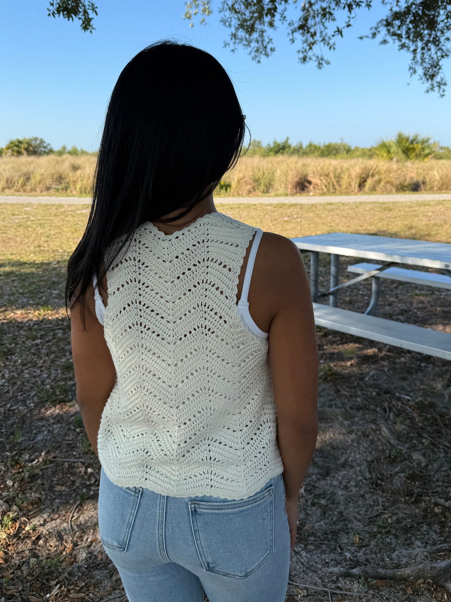 Woman wearing a white sleeveless crochet top and blue jeans standing outdoors with a picnic table and grass in the background.