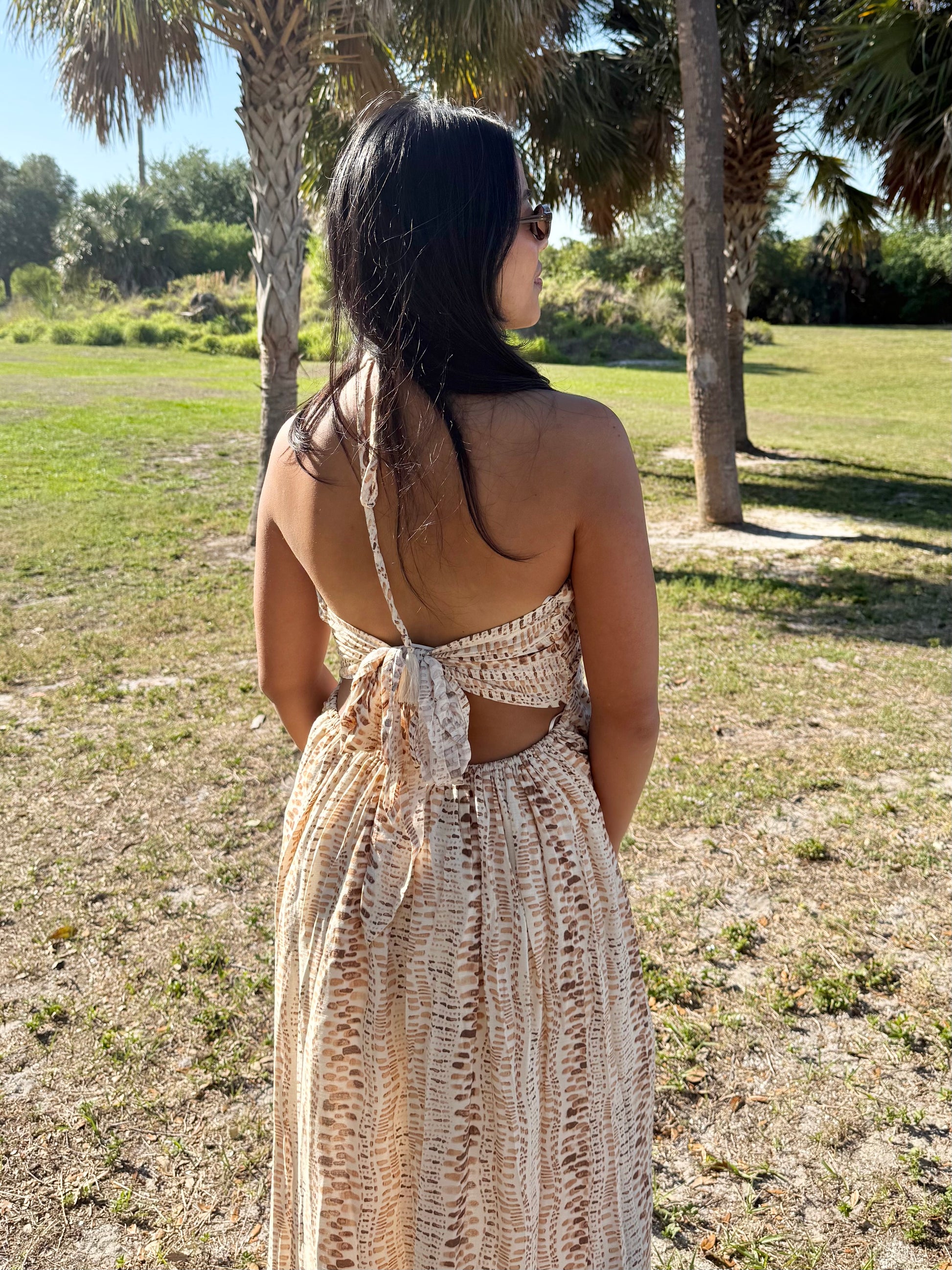 Woman in a long, open-back dress standing in a grassy area with trees.