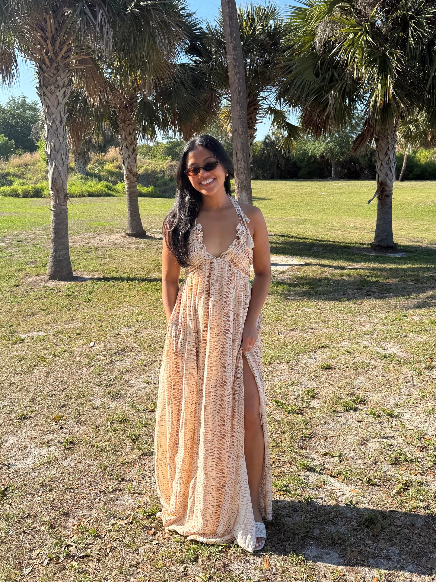 Woman in a long, patterned dress standing in a grassy area with palm trees.