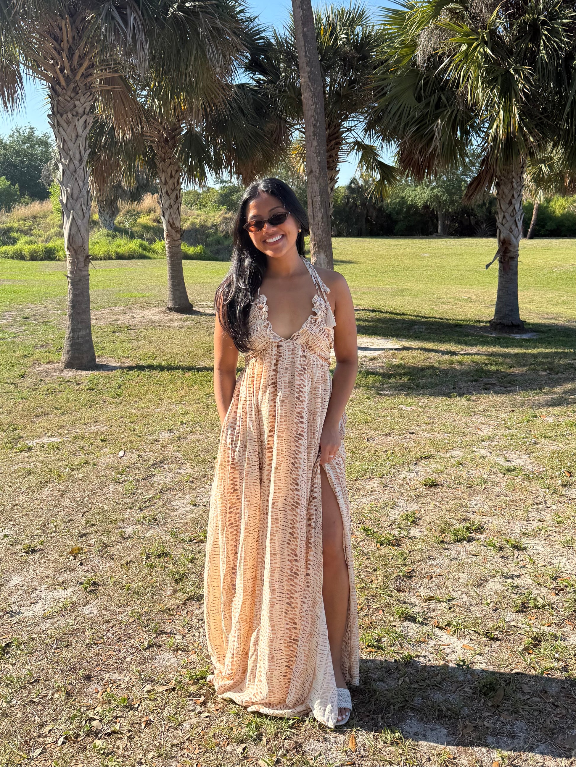 Woman in a long, patterned dress standing in a grassy area with palm trees.
