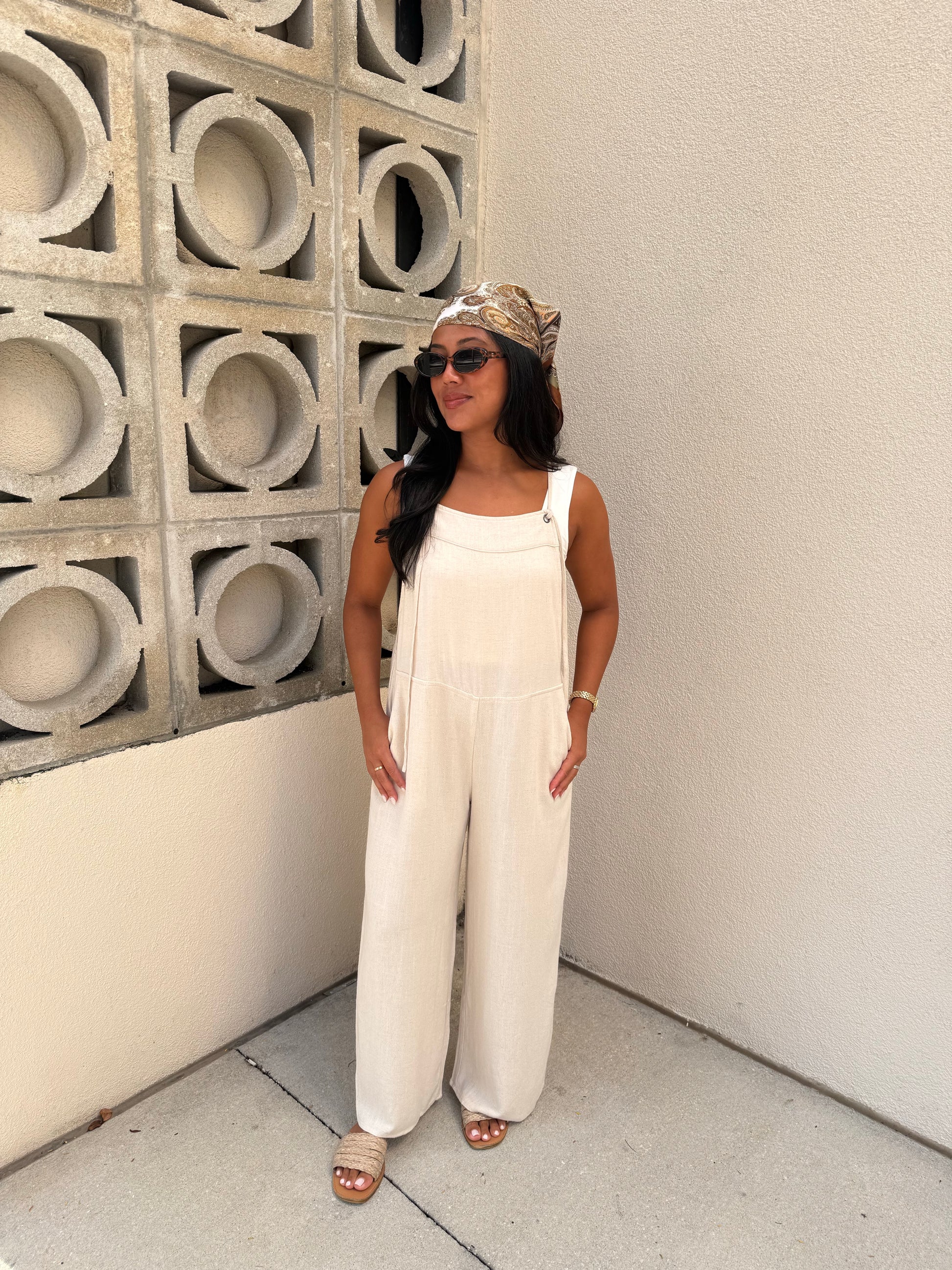 A woman wearing a white sleeveless wide-leg jumpsuit, a patterned headwrap, sunglasses, and flat sandals, standing in a corner with a concrete block wall and textured beige wall.