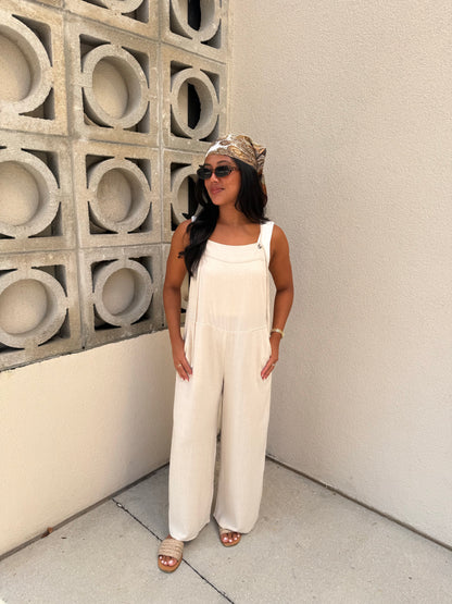 A woman wearing a white sleeveless wide-leg jumpsuit, a patterned headwrap, sunglasses, and flat sandals, standing in a corner with a concrete block wall and textured beige wall.