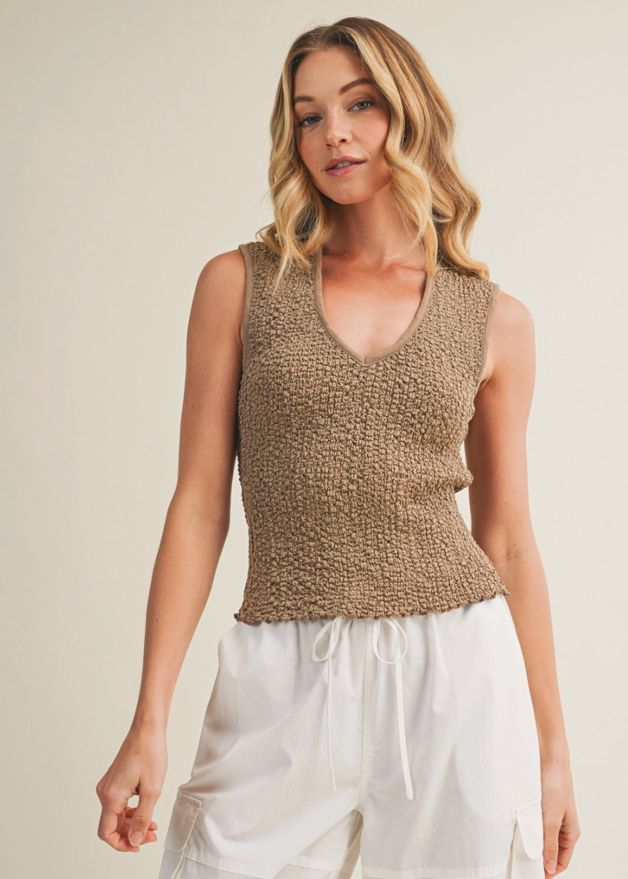 Woman wearing a brown knitted sleeveless top and white pants against a beige background