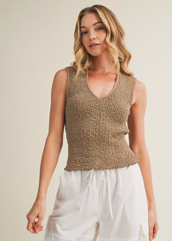 Woman wearing a brown knitted sleeveless top and white pants against a beige background