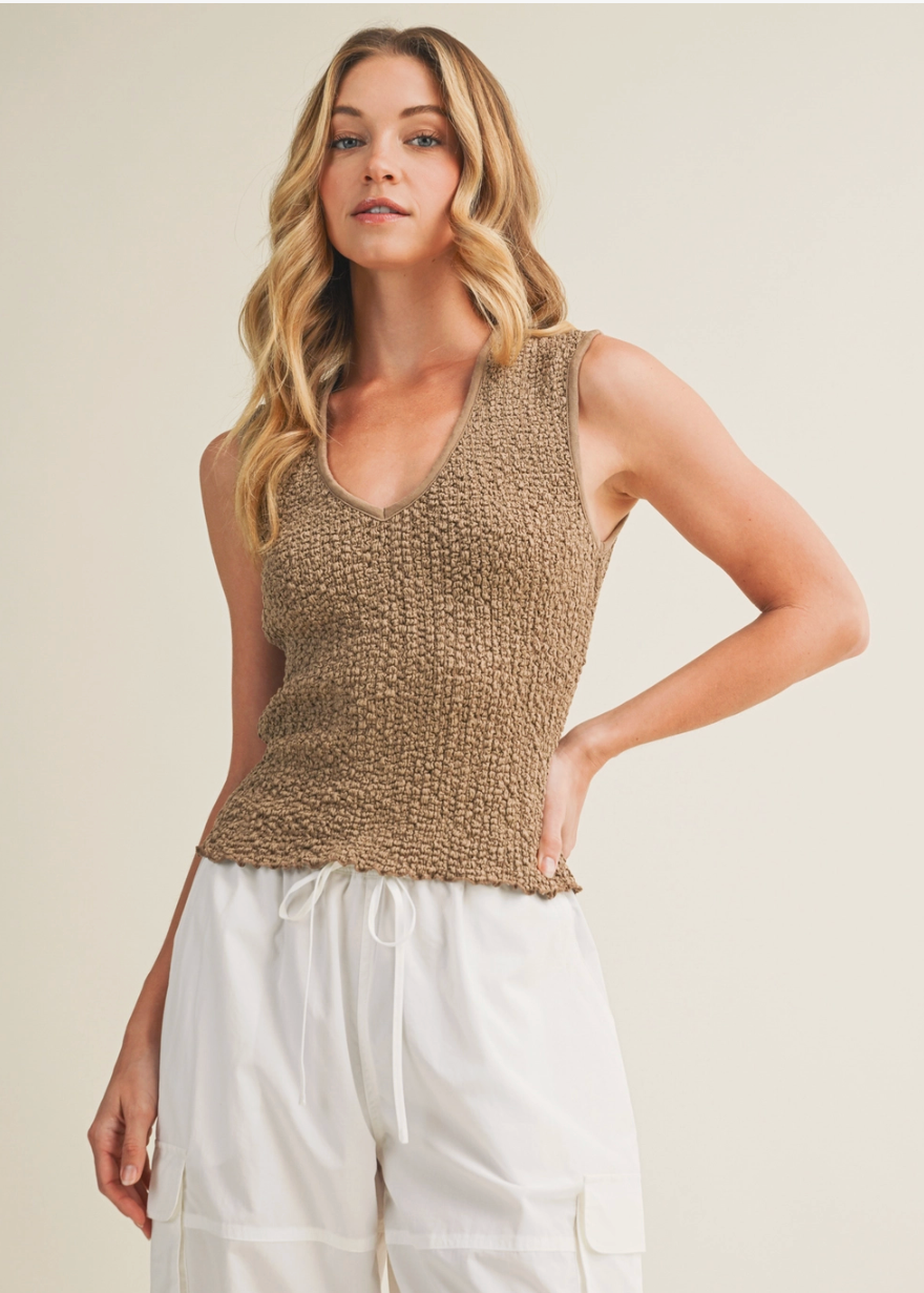 Woman wearing a brown knitted sleeveless top and white pants against a beige background