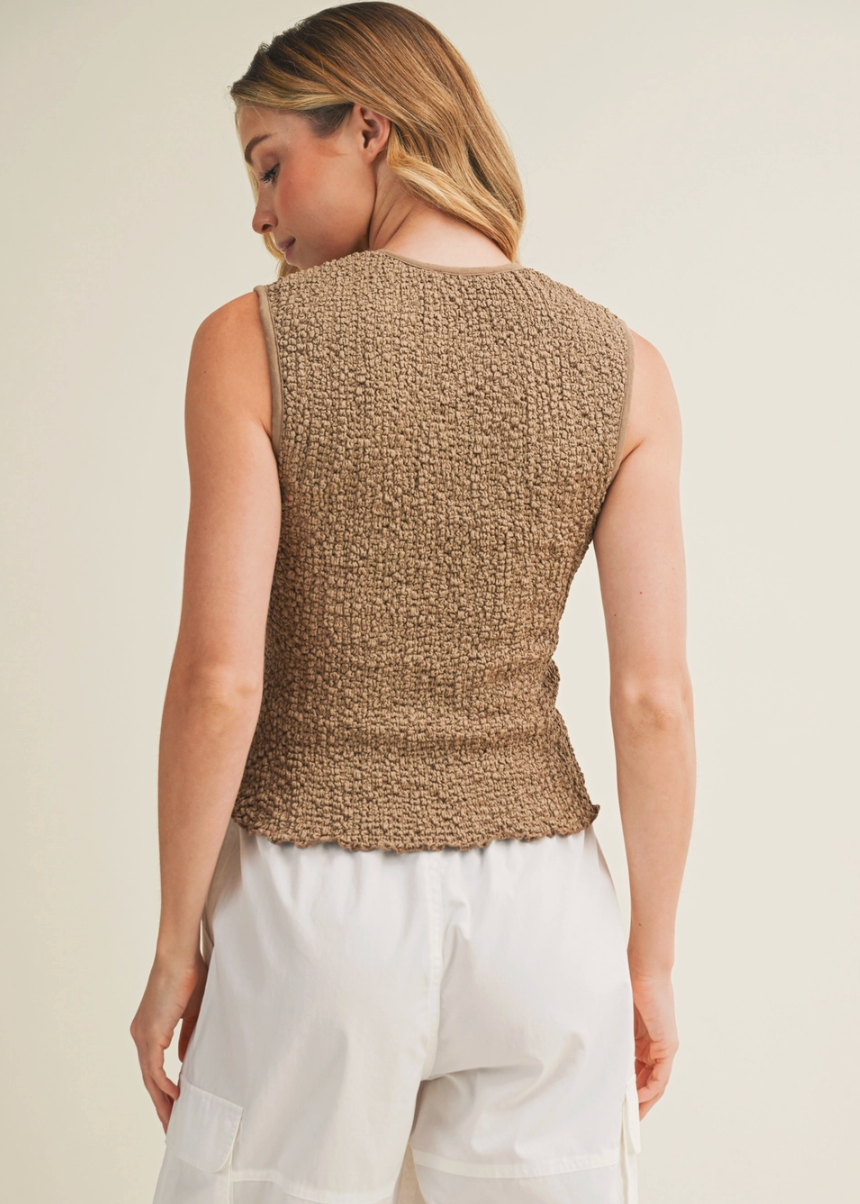 Woman wearing a beige knitted sleeveless top and white pants on a plain background
