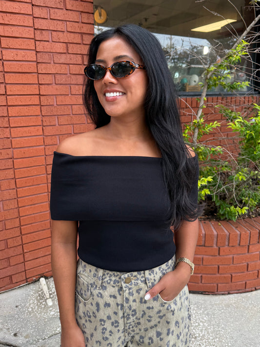 A woman wearing a black off-the-shoulder ribbed top and leopard-print pants, standing outdoors in front of a red brick wall.
