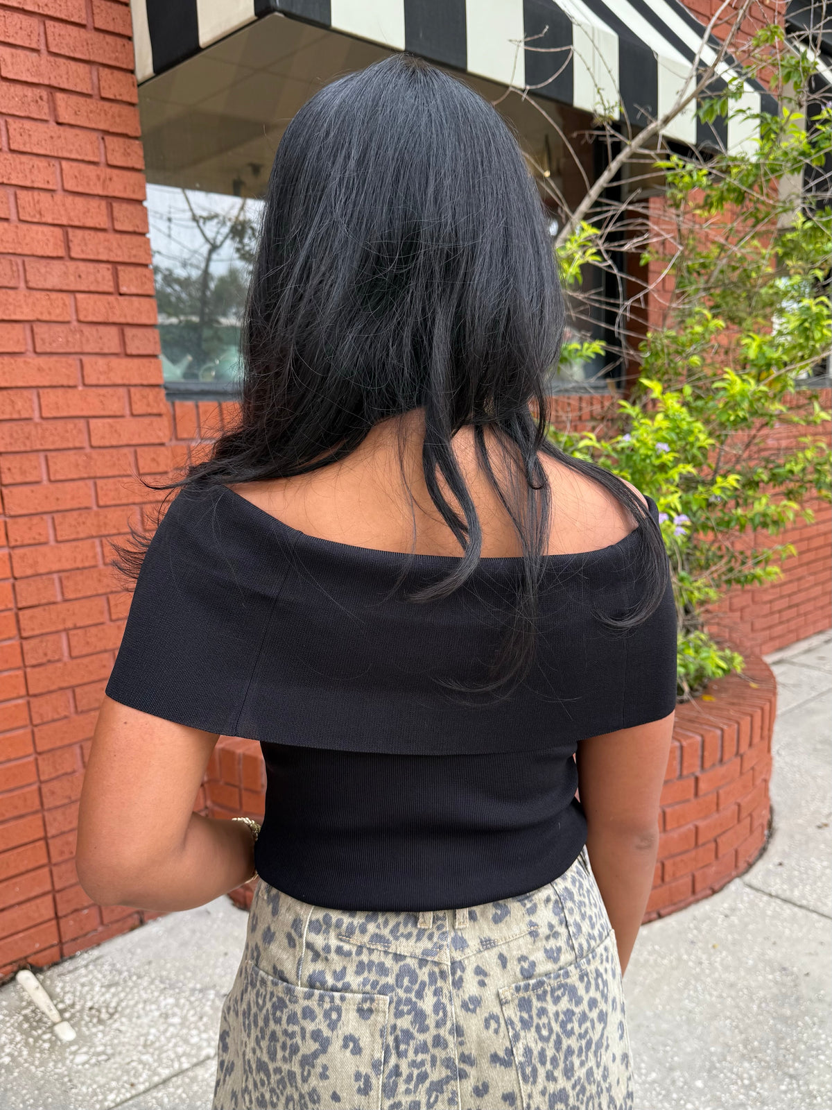 A woman viewed from behind wearing a black off-the-shoulder short-sleeve top and a beige leopard-print pant, standing outdoors next to a red brick wall.