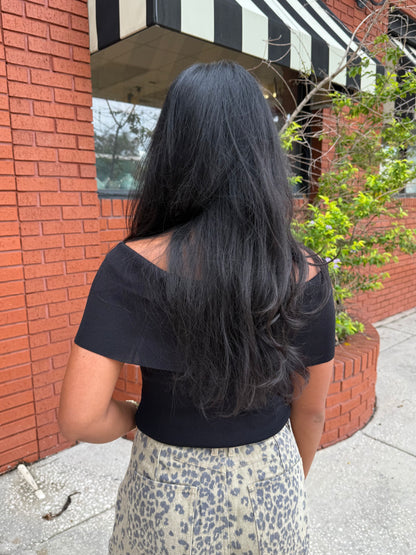 A woman viewed from behind wearing a black off-the-shoulder short-sleeve crop top and a beige leopard-print pant, standing outdoors in front of a red brick building.