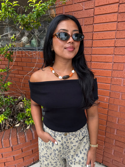 A woman with long black hair wearing a black off-the-shoulder top, leopard-print pants, and sunglasses, standing in front of a red brick wall.