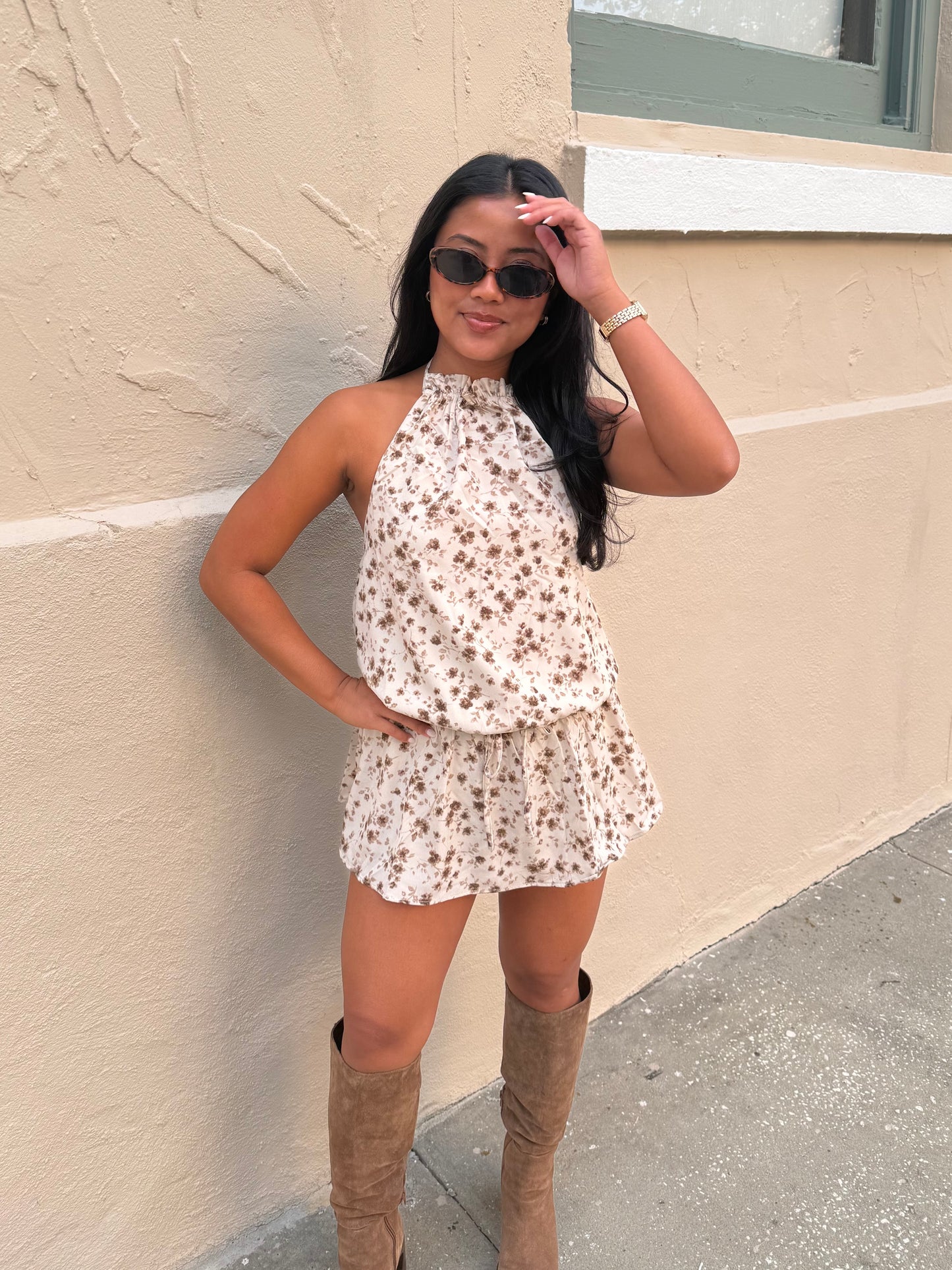 A woman wearing a sleeveless cream halter romper with a brown floral print and tan knee-high boots, posing against a beige stucco wall.