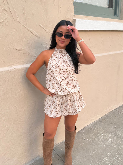 A woman wearing a sleeveless cream halter romper with a brown floral print and tan knee-high boots, posing against a beige stucco wall.