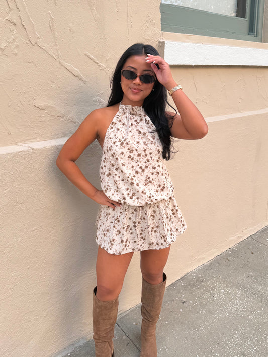 A woman wearing a sleeveless cream halter romper with a brown floral print and tan knee-high boots, posing against a beige stucco wall.