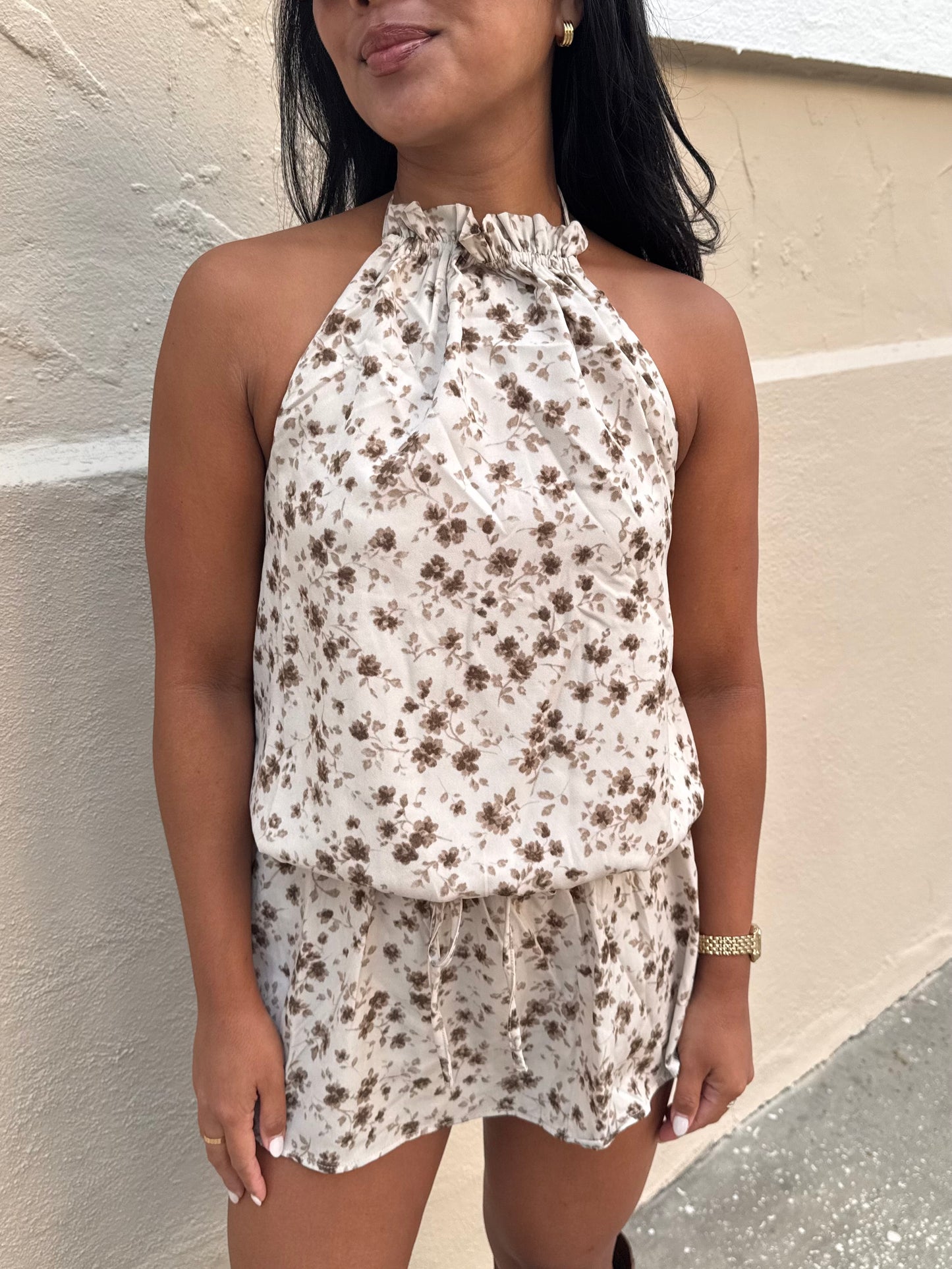A woman wearing a sleeveless halter-neck romper with a brown floral print on a cream background, standing against a beige stucco wall.