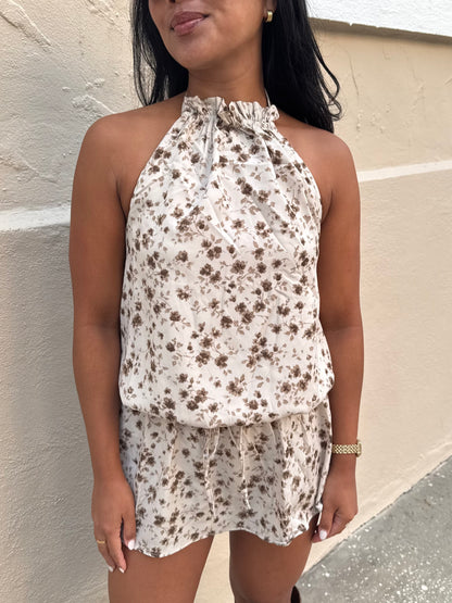 A woman wearing a sleeveless halter-neck romper with a brown floral print on a cream background, standing against a beige stucco wall.