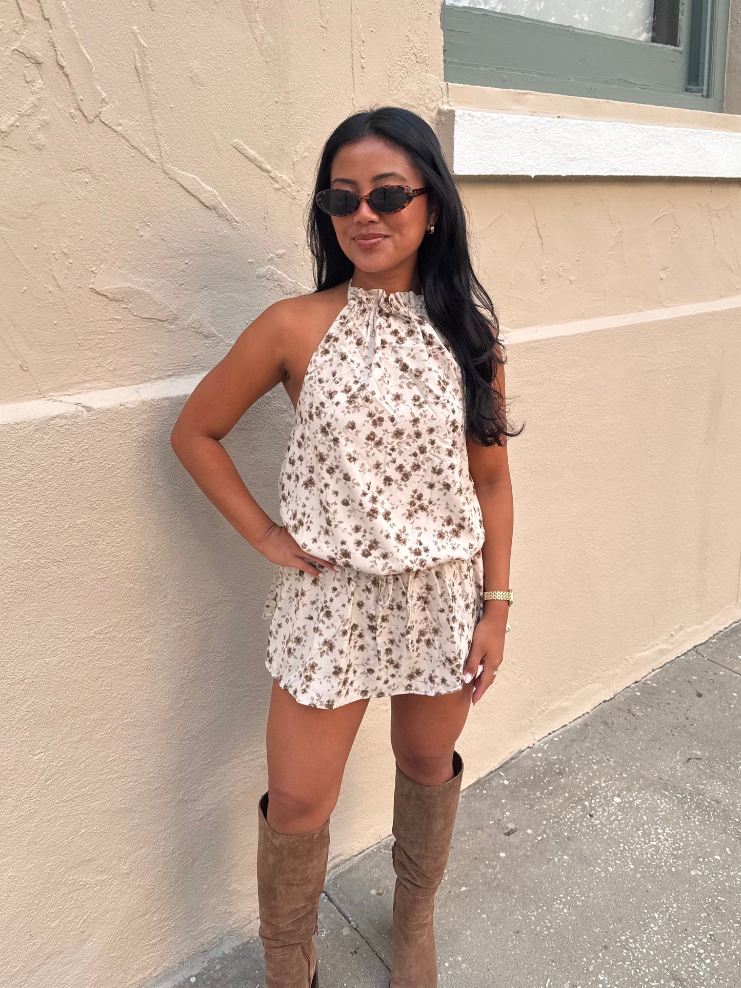 A woman wearing a beige and brown floral halter top and matching shorts, styled with brown knee-high boots and black sunglasses, posing against a beige stucco wall.