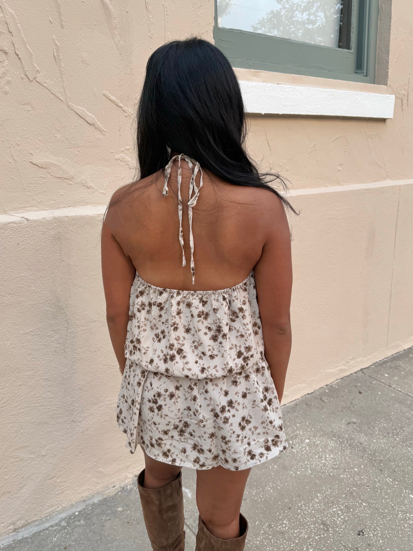 A woman viewed from behind wearing a cream halter mini dress with a brown floral pattern and tie-back straps, paired with brown knee-high boots, standing against a beige stucco wall.