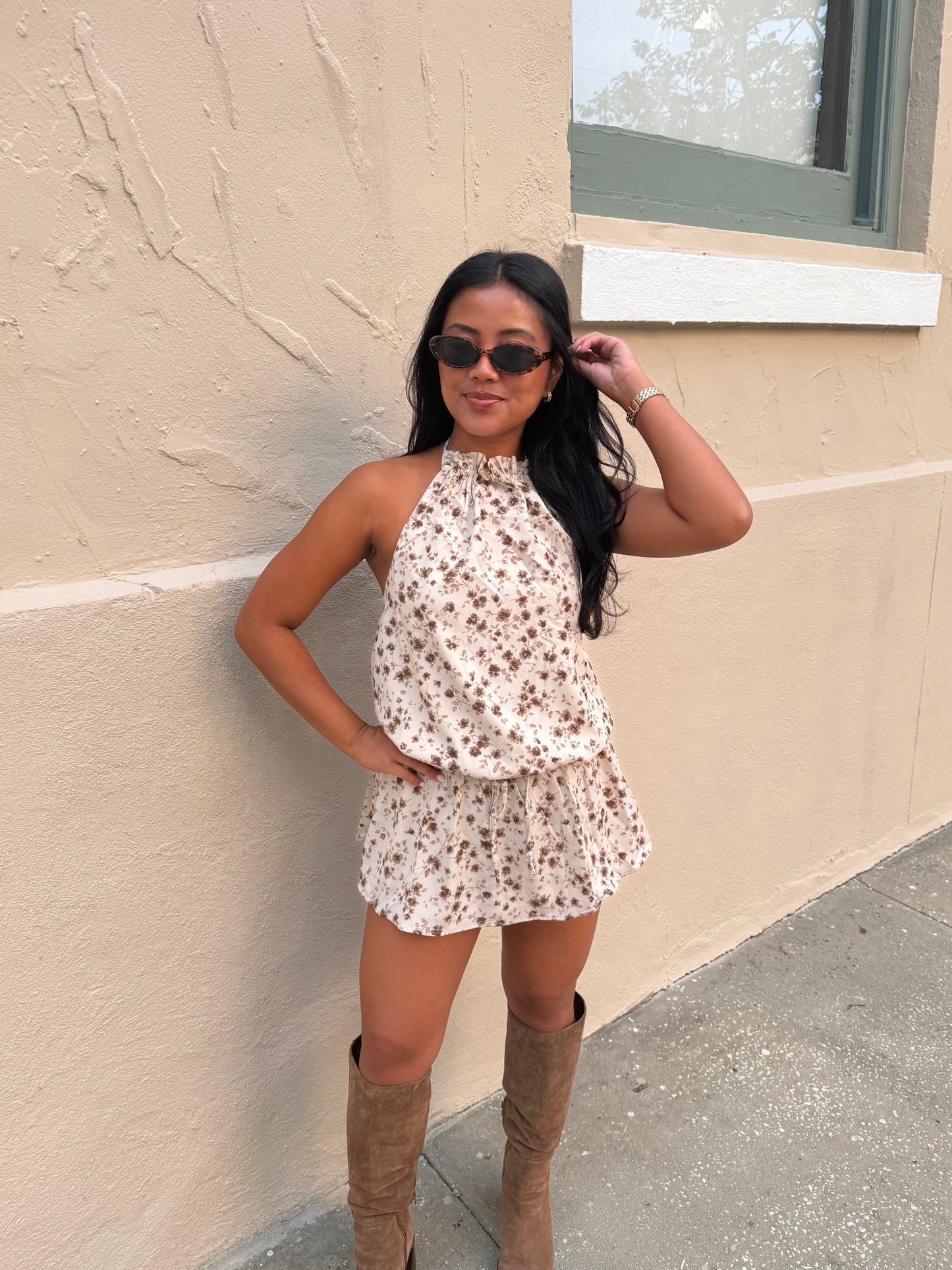 A woman wearing a sleeveless cream halter romper with a brown floral pattern and brown knee-high boots, posing against a beige stucco wall.