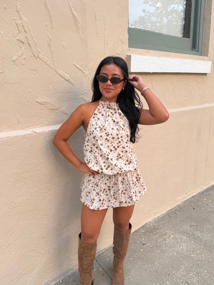 A woman wearing a sleeveless cream halter romper with a brown floral pattern and brown knee-high boots, posing against a beige stucco wall.