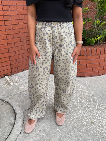 A person wearing loose, wide-leg pants with a beige and gray leopard print pattern, paired with a black top and pink flats, standing outdoors against a red brick wall.